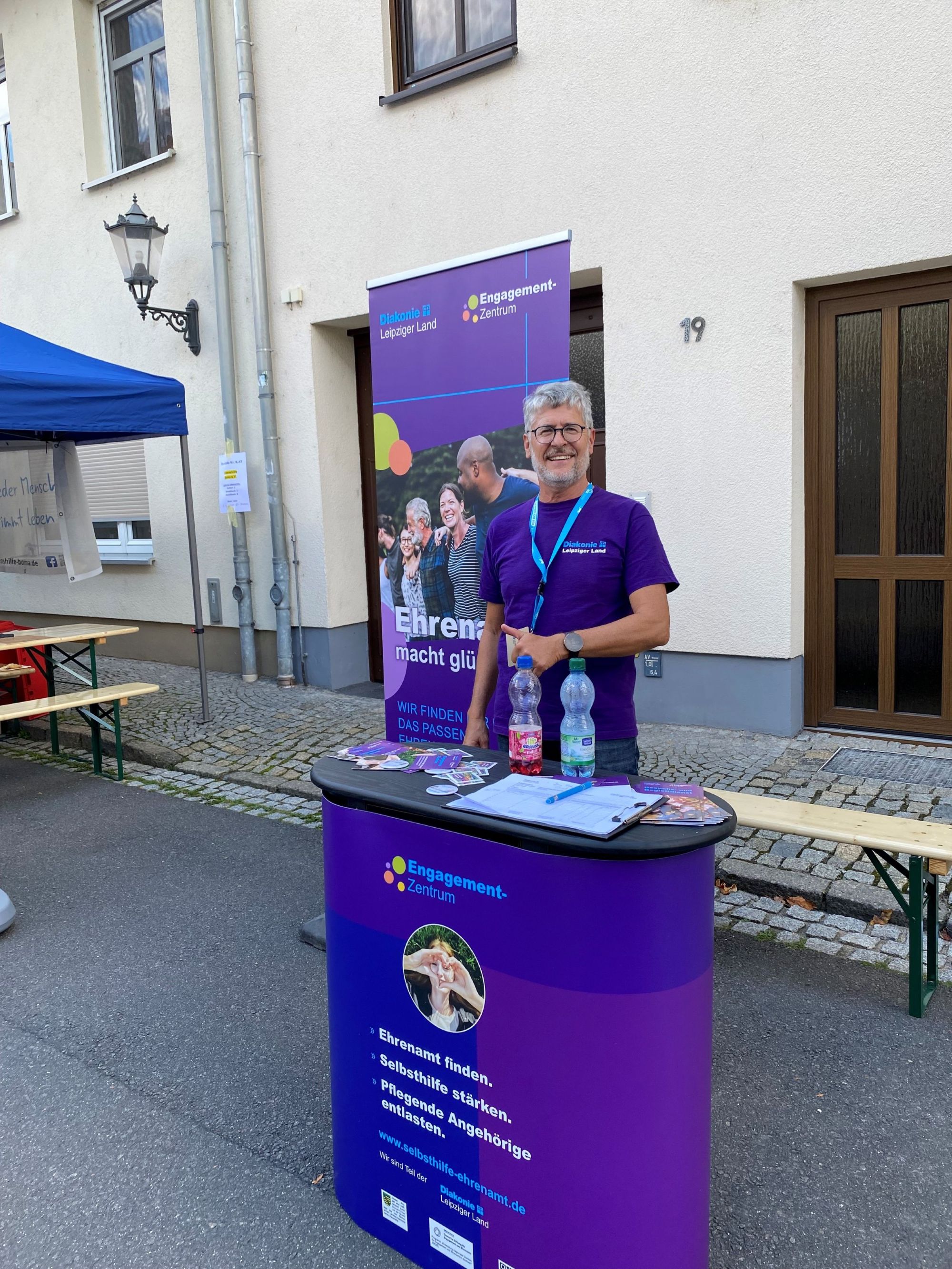Auf dem Foto ist der Engagement-Scout der Diakonie auf dem Stadtfest in Borna zu sehen. Er steht lächelnd hinter einer lila Messetheke und blickt in die Kamera. Hinter ihm steht ein lila Roll-Up mit der Aufschrift "Ehrenamt macht glücklich."